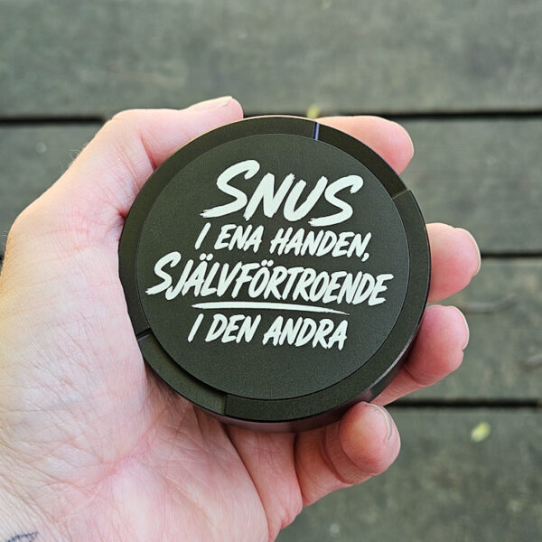 Svart snusdosa med texten Snus i ena handen självförtroende i den andra